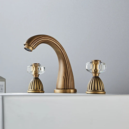 An Antique Bronze Double Handle Bathroom Faucet by Tiqui stands on a white countertop. It has two crystal-like handles on either side, also featuring elegant detailing, giving the entire fixture an antique and luxurious appearance.