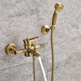 Ellen's Wasser™ Antique Brass Shower Faucet with Bathtub Faucet is mounted on a textured stone wall. The setup features a shower faucet with a lever handle, a diverter for switching water flow, and a detachable handheld showerhead with a flexible hose. Water is flowing from the faucet.