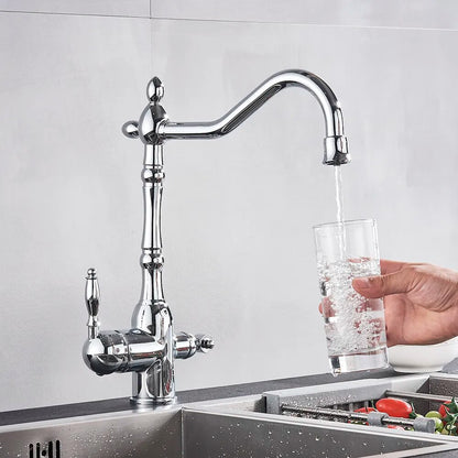 A modern Uythner chrome kitchen faucet with a filtered water tap releases a steady stream of water into a stainless steel sink filled with various vegetables. A ceramic bowl with an intricate pattern sits on the countertop, and the background is a minimalist gray wall.