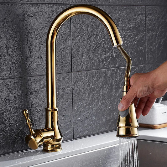 A hand is holding the nozzle of the Tiqui Pull Out Spout Single Handle Kitchen Faucet, which features a shiny gold finish and a pull-down sprayer, mounted on a stainless steel sink. Water flows from the kitchen faucet against a backdrop of dark gray tiled wall.