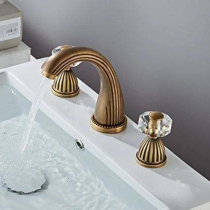 An Antique Bronze Double Handle Bathroom Faucet by Tiqui stands on a white countertop. It has two crystal-like handles on either side, also featuring elegant detailing, giving the entire fixture an antique and luxurious appearance.