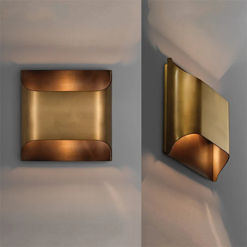 The Rame Modern Copper Wall Lights by Morsale are wall-mounted fixtures with a sleek geometric design, made from polished brass. With a square front and an angled side view, these lamps cast a warm glow using LED bulbs against a gray backdrop.