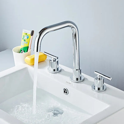 A Double Handle Solid Brass Deck Mounted Bathroom Faucet by Tiqui in a shiny gold finish stands out against a white marble backsplash. The faucet features a high-arched spout with two matching gold handles on either side, all elegantly positioned on a white countertop to create a modern and sophisticated design.