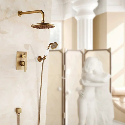 A luxurious bathroom features the Rozin Antique Brass Shower System with handheld shower mounted on a marble wall. In the background, a blurred decorative sculpture of two embracing figures and a sunlight-lit column add elegance to the room, while a matching gold bathroom sink faucet complements the opulent design.