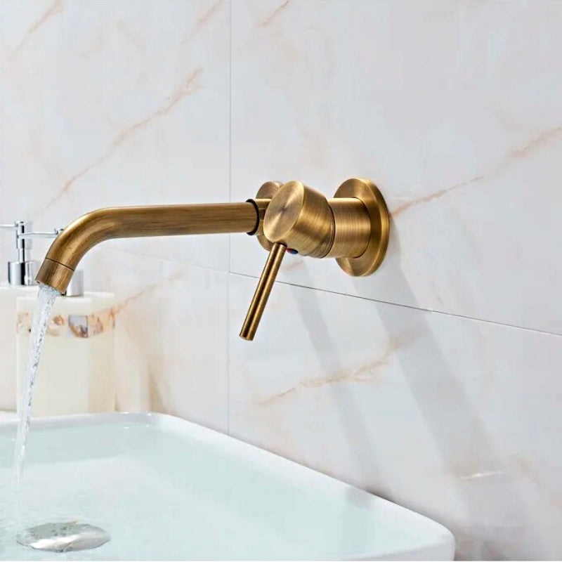 The Rozin Antique Brass Wall Mounted Bathroom Faucet pours water into a deep white sink, set against beige marble tiles. The steady, clear stream and minimalist design impart a modern aesthetic to the setup.