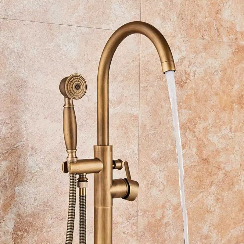 A Rozin Antique Brass Floor Mounted Bathtub Filler Shower System, featuring an arched spout and a handheld shower head connected by a flexible hose, is installed on a beige marble-tiled wall. Water flows elegantly from the spout into an unseen bathtub or sink.