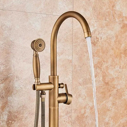 A Rozin Antique Brass Floor Mounted Bathtub Filler Shower System, featuring an arched spout and a handheld shower head connected by a flexible hose, is installed on a beige marble-tiled wall. Water flows elegantly from the spout into an unseen bathtub or sink.