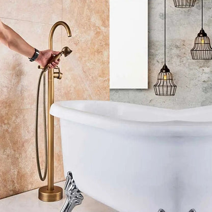 A Rozin Antique Brass Floor Mounted Bathtub Filler Shower System, featuring an arched spout and a handheld shower head connected by a flexible hose, is installed on a beige marble-tiled wall. Water flows elegantly from the spout into an unseen bathtub or sink.