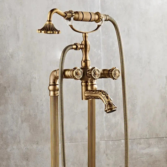 The Antique Brass Floor Standing Bathtub Faucet Tub Filler by Allfixture is a stunning vintage piece. It features an elegantly designed handheld shower head and dual temperature control knobs. The fixture boasts an antique brass finish with ornate detailing, curved handles, and flexible hoses, perfectly set against a neutral-toned tiled wall.