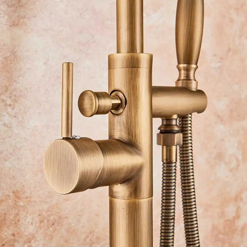 A Rozin Antique Brass Floor Mounted Bathtub Filler Shower System, featuring an arched spout and a handheld shower head connected by a flexible hose, is installed on a beige marble-tiled wall. Water flows elegantly from the spout into an unseen bathtub or sink.