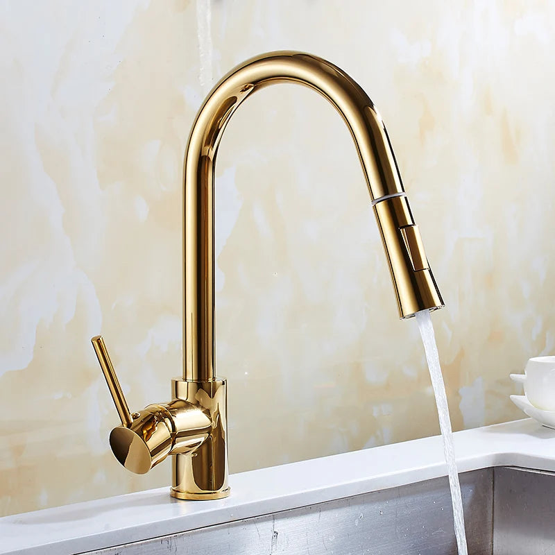 A hand holding the detachable spray head of Tiqui's Single Handle Pull Out Kitchen Faucet, directing the water stream into a stainless steel sink. The sleek, gold-colored faucet features a single lever handle for controlling water flow and temperature. The background shows a marble-like wall.