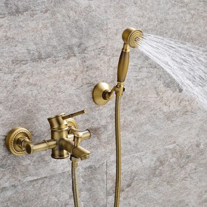 Ellen's Wasser™ Antique Brass Shower Faucet with Bathtub Faucet is mounted on a textured stone wall. The setup features a shower faucet with a lever handle, a diverter for switching water flow, and a detachable handheld showerhead with a flexible hose. Water is flowing from the faucet.