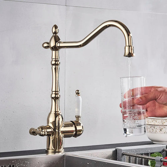 A polished gold kitchen faucet with a filtered water tap and vintage design by Uythner is mounted on a countertop. Water is flowing from the faucet into a clear glass being held by a person's hand. A small dish and green herbs are visible in the background.