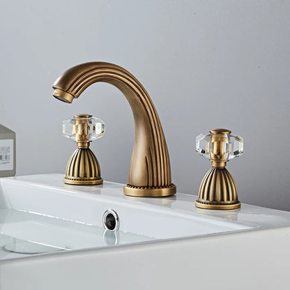 An Antique Bronze Double Handle Bathroom Faucet by Tiqui stands on a white countertop. It has two crystal-like handles on either side, also featuring elegant detailing, giving the entire fixture an antique and luxurious appearance.