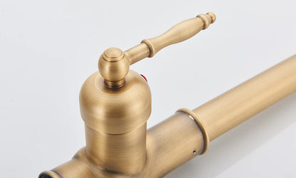 A close-up of the Rozin Antique Brass Pull-Down Spout Kitchen Faucet with a high arc and a pull-down sprayer. A hand is shown holding the sprayer, with water flowing into a stainless steel sink. A white curtain and a white bowl with purple flowers are visible in the background, enhancing the kitchen's charm.
