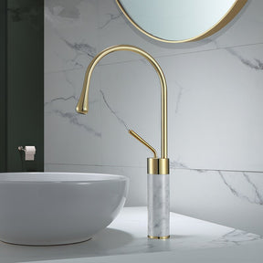 A sleek, modern bathroom sink setup featuring the Tiqui Solid Brass & Marble Bathroom Basin Faucet, showcasing a tall, curved gold design with a matching handle. Below the faucet is a stunning black marbled bowl sink. An elegant shower head complements the light grey tiles in the background, which have subtle marble veining.