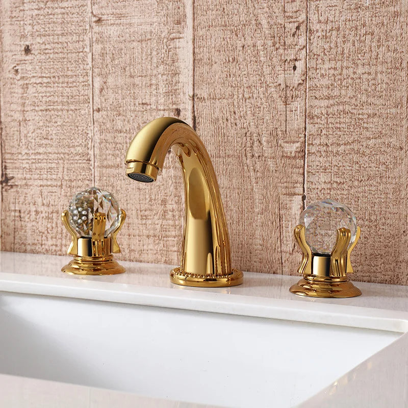 The AllFixture Luxurious Brass and Marble Bathroom Faucet - Widespread 3-Hole Gold and Marble Sink Mixer Tap features an elegant design with a gold-plated finish and brown marble accents. The set includes a curved spout flanked by two handles, all mounted on a clean, sleek sink. In the background, you'll find a gray wall and neatly folded beige towels completing the sophisticated look.