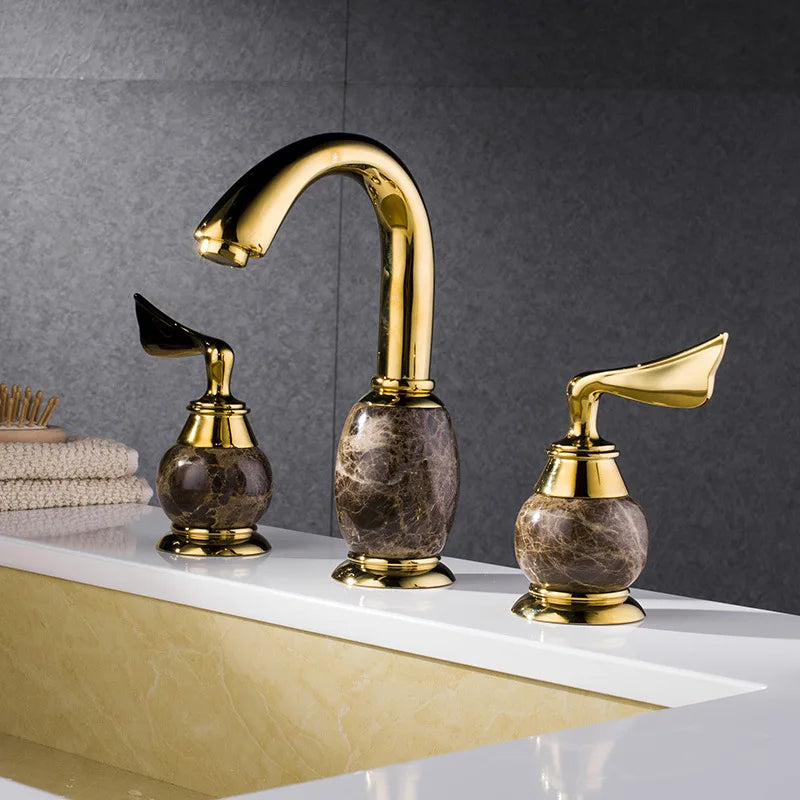 The AllFixture Luxurious Brass and Marble Bathroom Faucet - Widespread 3-Hole Gold and Marble Sink Mixer Tap features an elegant design with a gold-plated finish and brown marble accents. The set includes a curved spout flanked by two handles, all mounted on a clean, sleek sink. In the background, you'll find a gray wall and neatly folded beige towels completing the sophisticated look.