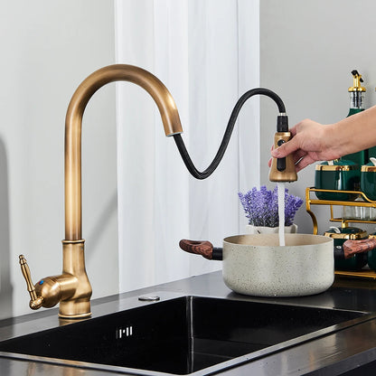 A close-up of the Rozin Antique Brass Pull-Down Spout Kitchen Faucet with a high arc and a pull-down sprayer. A hand is shown holding the sprayer, with water flowing into a stainless steel sink. A white curtain and a white bowl with purple flowers are visible in the background, enhancing the kitchen's charm.
