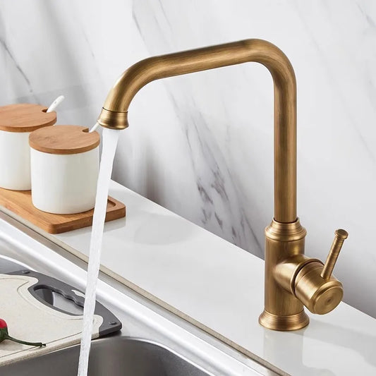 A modern Solid Brass Antique Kitchen Sink Faucet by Tiqui with a brushed bronze finish is shown mounted on a white countertop. Water flows from the faucet, and two white containers with wooden lids are placed in the background. Part of a stainless steel sink is visible in the foreground.