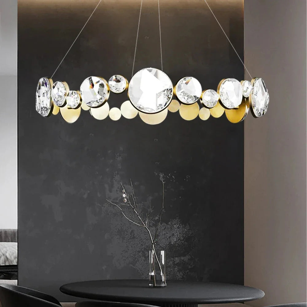 A Morsale Diamante Crystal Ceiling Light Fixture, featuring circular glass and metal elements, hangs above a dark round table. The setting includes a textured gray wall backdrop and a minimalistic vase adorned with bare branches on the table.