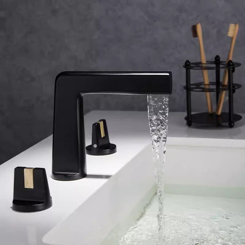 A sleek, modern Solid Brass Dual Handle Bathroom Faucet by Tiqui, featuring a rectangular spout in a brass finish, cascades water into a white sink. Flanked by two matching handles on each side, the minimalist design stands out against a dark gray backdrop. Complementing this piece are equally elegant Tiqui shower faucets to maintain a cohesive aesthetic.