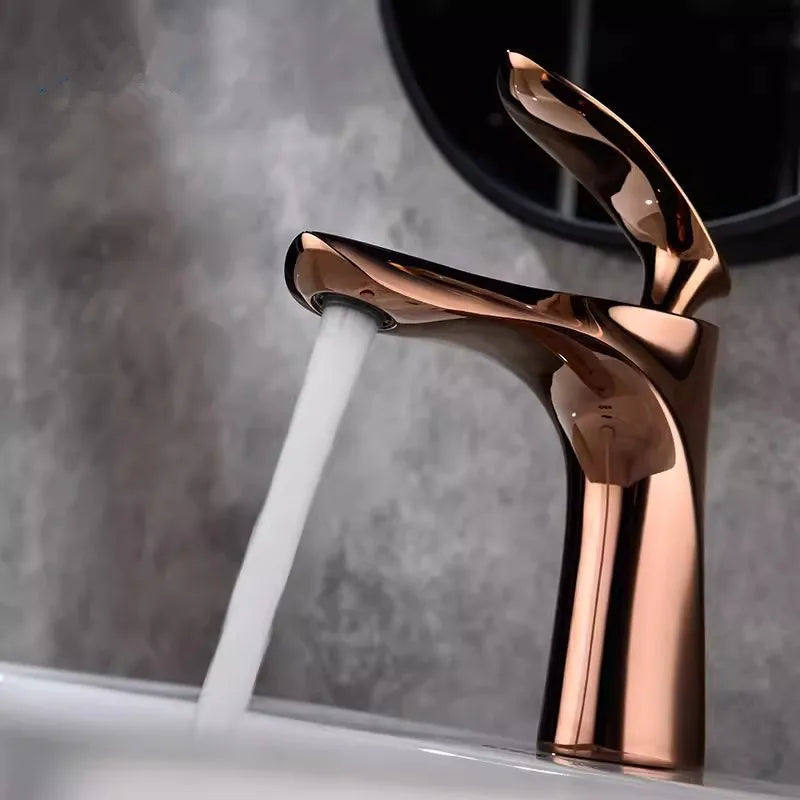 A close-up view of the Allfixture Rose Gold Bathroom Faucet – Deck Mounted Basin Sink Mixer showcases water gracefully flowing into a pristine white sink. The background features a gray textured wall. This faucet boasts a contemporary design with smooth curves and a polished finish, epitomizing modern bathroom designs.