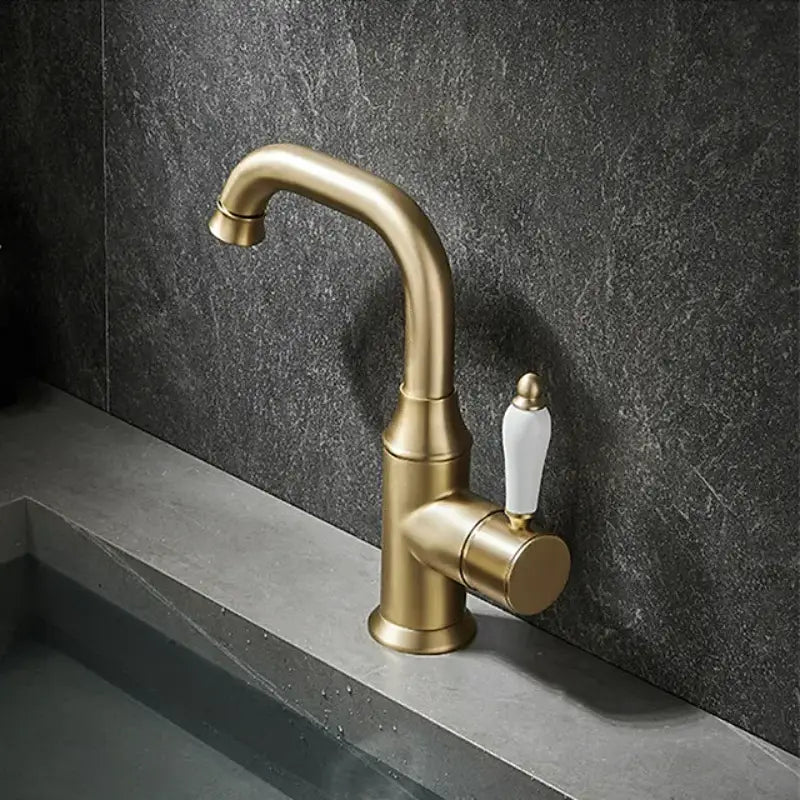 The Tiqui™ Single Handle Bathroom Sink Faucet, featuring a brass finish and white handle, is installed over a gray countertop, pouring a stream of water into the sink. The background features a dark textured wall and a small vase with white flowers.