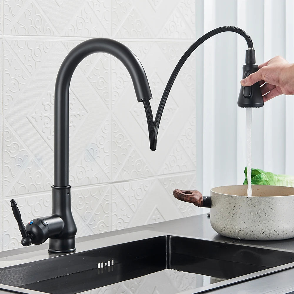 A Rozin Matte Black Pull-Down Kitchen Faucet with a high arc neck and single lever handle is seen in action, streaming water into a stainless steel sink. In the background, there is a leafy green vegetable and a cutting board on the countertop. The backsplash features a geometric design reminiscent of modern shower head patterns.