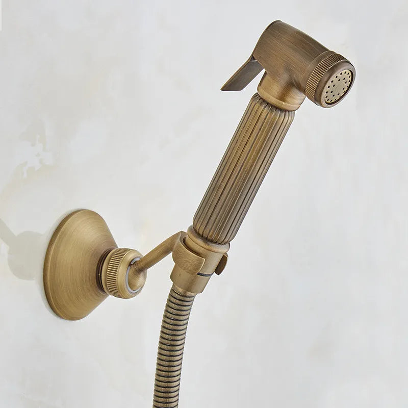 The Allfixture Antique Brass Wall Mounted Bidet Faucet Hand shower boasts a vintage-style design with a ribbed handle and a brushed bronze finish. Mounted on the wall, it is crafted from brass material and features a small nozzle along with a thumb-controlled lever, all attached to a flexible metal hose.