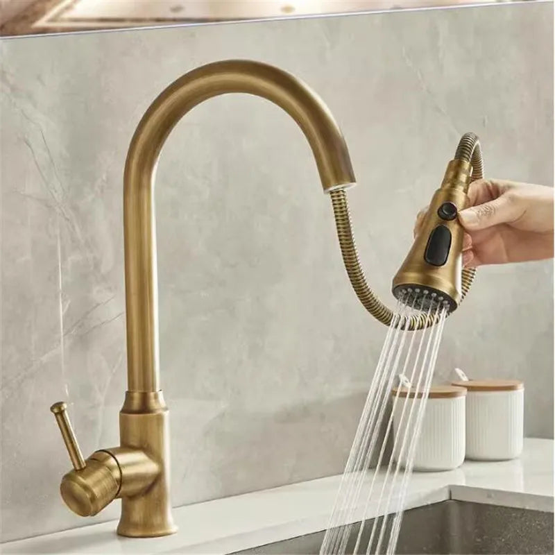 A hand grips the pull-down spout of the AllFixture Maison Antique Brass Kitchen Faucet, featuring a curved design and single handle, as water streams into the sink. Two white containers are visible on the countertop in the background.