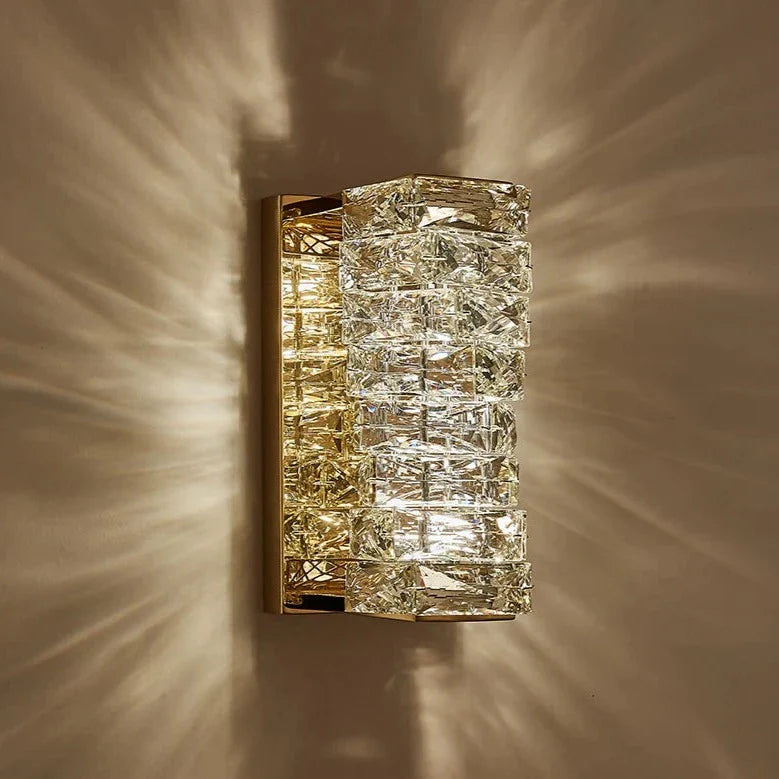 The Bacci Crystal Wall Sconces by Morsale.com feature a gold-finish and intricate crystal glass detailing, supported by a brass frame, illuminating walls with a warm and decorative pattern of light and shadows. These modern sconces are elegant and luxurious, seamlessly blending artistic elements with classic opulence.
