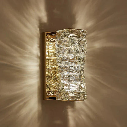 The Bacci Crystal Wall Sconces by Morsale.com feature a gold-finish and intricate crystal glass detailing, supported by a brass frame, illuminating walls with a warm and decorative pattern of light and shadows. These modern sconces are elegant and luxurious, seamlessly blending artistic elements with classic opulence.