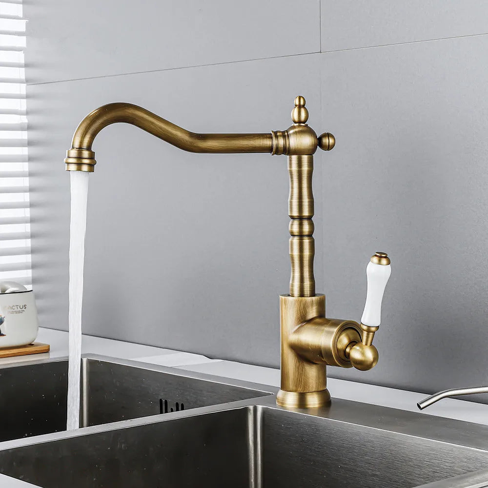 An Allfixture Single Handle Vintage Kitchen Sink Faucet is elegantly perched on a stainless steel sink. This deck-mounted brass faucet features a ceramic plate spool and dispenses water with ease, while its handle boasts a white accent. The backdrop showcases a gray tiled wall and a small potted plant.