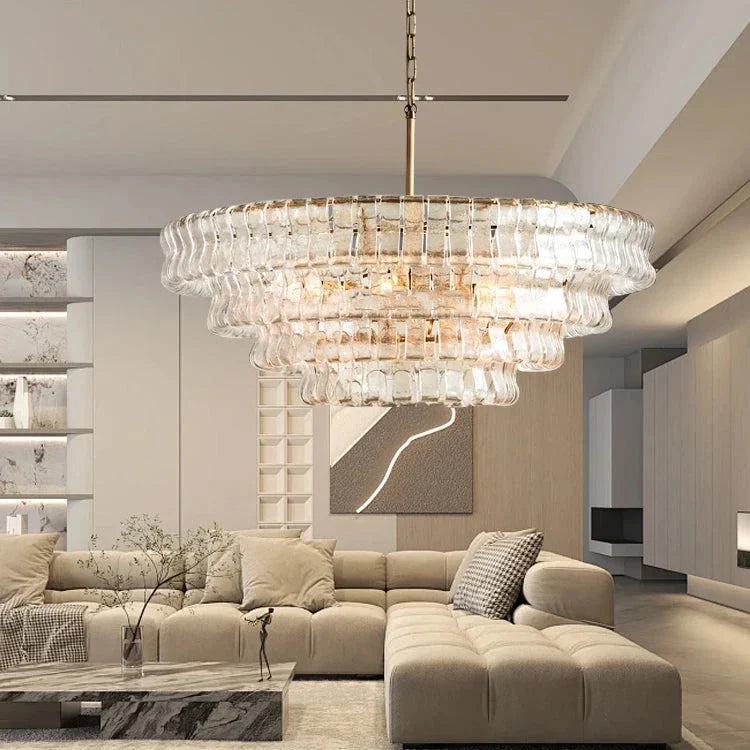 A modern living room featuring the Riviera Vintage French Glass Chandelier by Morsale, which is handcrafted with a tiered glass design. Below is a beige sectional sofa and a marble coffee table. The room showcases a minimalist aesthetic with light-colored walls and a decorative panel, capturing the essence of modern style.