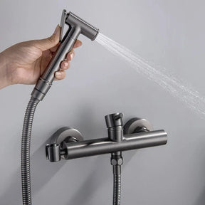 The Tiqui Cold And Hot Water Wall Mounted Bidet Faucet, featuring a sleek, gold-toned finish with a cylindrical control handle and flexible hose, boasts a minimalist yet contemporary design. This modern fixture, reminiscent of premium bathroom faucets, is mounted on a gray wall for an elegant look.