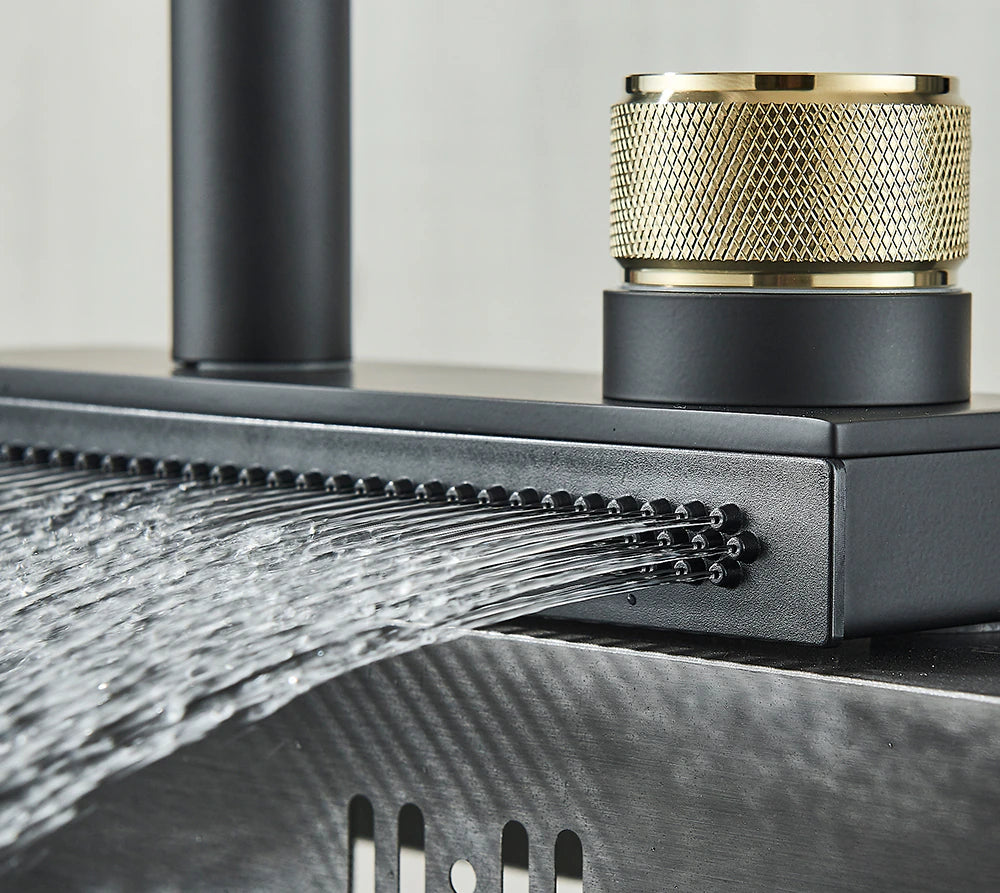 The kitchen sink is graced by the modern Rainfall Pull Out Spout Kitchen Faucet from Rozin, featuring a high-arc spout in a sleek matte black finish. Water cascades vertically from the front of the faucet in a flat stream. A gold-colored handle is prominently situated to the right of the base, while a potted plant peeks into view on the left.