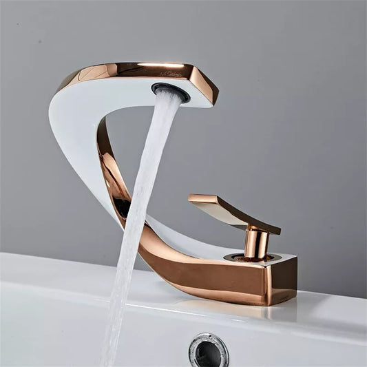 A modern White & Rose Gold Bathroom Sink Faucet by allfixture, featuring a curved design, is dispensing water into a white sink against a gray wall. The faucet, resembling high-end bathroom faucets, has a sleek handle for controlling the water flow.