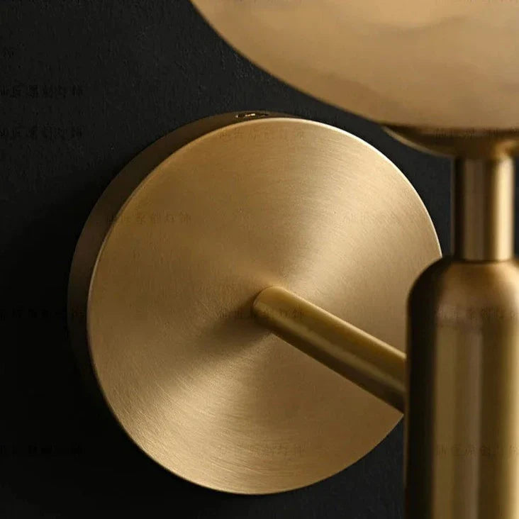 A Natural Marble Sphere Wall Sconce by Morsale.com with a round, frosted glass globe lampshade. In the top view, the brass light source is mounted on a dark wall. In the bottom view, the sconce is installed on a light-colored wall next to a framed artwork and a potted plant with green leaves.