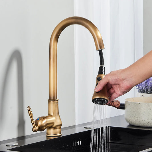 A close-up of the Rozin Antique Brass Pull-Down Spout Kitchen Faucet with a high arc and a pull-down sprayer. A hand is shown holding the sprayer, with water flowing into a stainless steel sink. A white curtain and a white bowl with purple flowers are visible in the background, enhancing the kitchen's charm.
