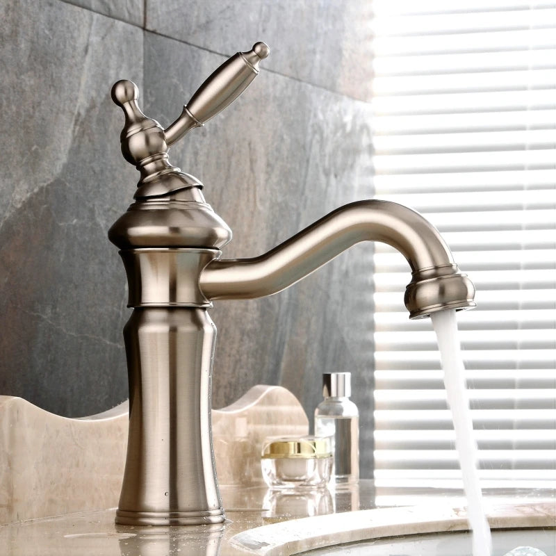 An Allfixture Solid Brass Single Handle Swivel Spout Bathroom Sink Faucet, featuring a vintage design, is deck mounted on a marble countertop, releasing a stream of water into the white sink below. Nearby, a small bottle of liquid soap and perfume bottle sit, with slatted blinds visible in the background.