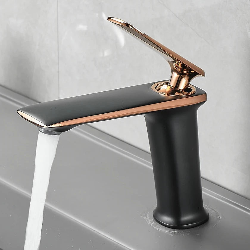 A sleek, modern Becola™ Brass Bathroom Faucet with a black and bronze finish is installed on a gray countertop. The faucet, branded by Becola, features a single lever handle with water flowing from the spout. The background showcases a light-colored, textured wall.