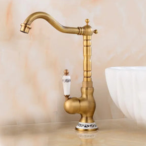 An Allfixture Tall Torneiras Bathroom Faucet – Single Handle, Hot and Cold Water Mixer, featuring a vintage-style brass finish with a white ceramic handle and decorative base, is installed next to a modern sink basin. Water flows from the faucet into the sink, set against a light beige background.