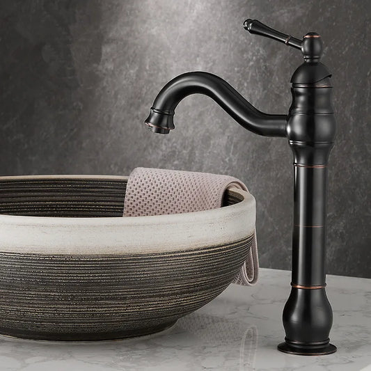 An Allfixture Deck Mounted Antique Brass Bathroom Faucet with an elegant curved spout and single handle stands beside a round, textured stone sink. Featuring a hot and cold water mixer, the tall, dark bronze faucet perfectly complements the folded beige towel draped over the sink edge against a dark gray marble wall and white marble countertop.