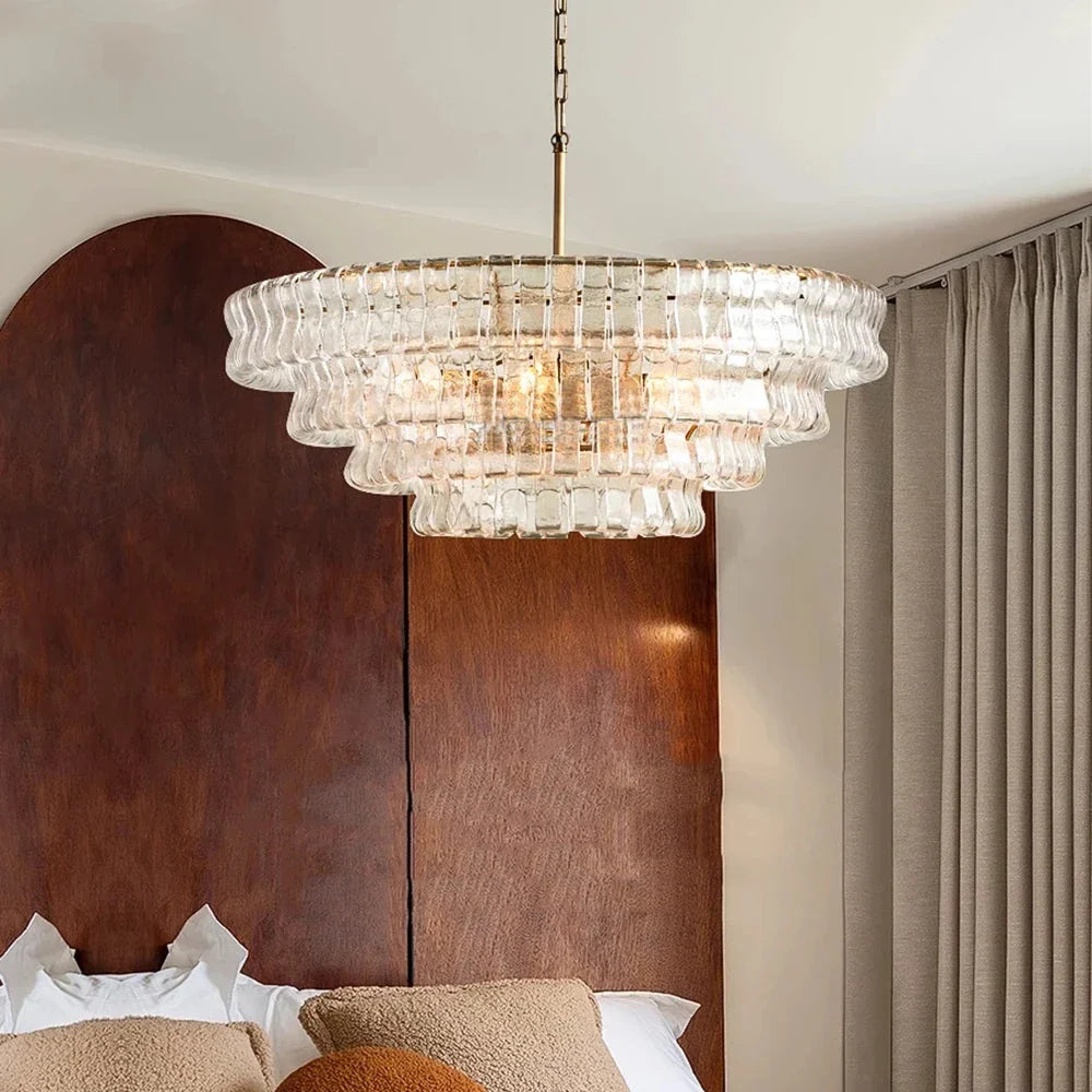 A contemporary bedroom features the Riviera Vintage French Glass Chandelier by Morsale, elegantly crafted above a bed decorated with white pillows and brown cushions. The bed is highlighted by a tall wooden headboard, paired beautifully with beige curtains cascading to the right.