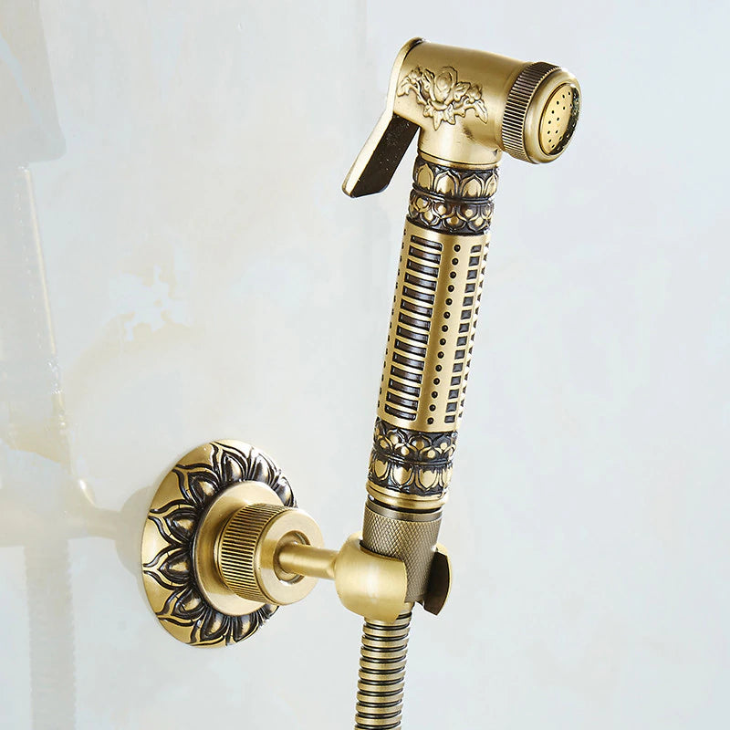 A decorative Wall Mounted Antique Bronze Toilet Sprayer Bidet Faucet from Tiqui set against a light background. The set includes an intricately designed faucet handle, a water spout with ornate detailing, and an adjustable handheld sprayer attached to a flexible hose. The design features floral and geometric patterns.