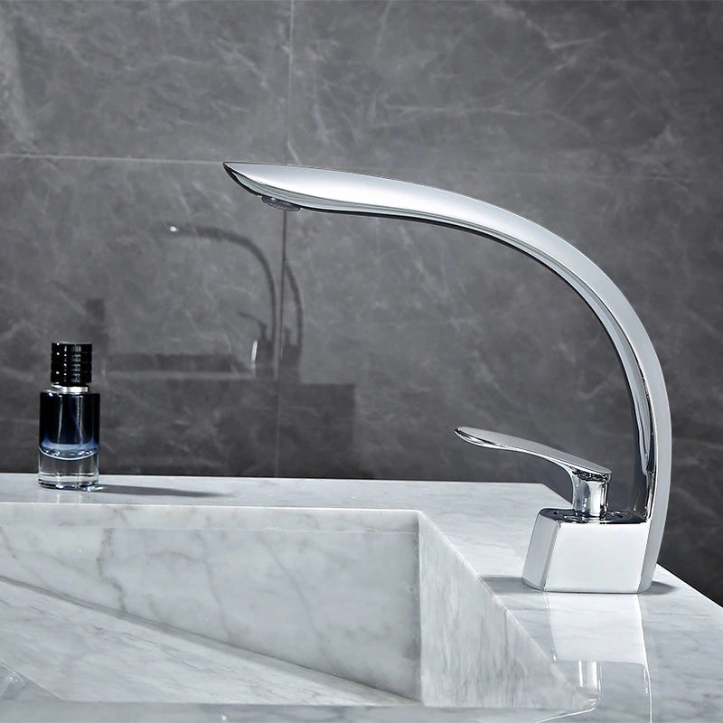 A sleek, modern Tiqui Wasser™ Single Handle Bathroom Sink Faucet with a curved spout and single lever handle is installed on a marble countertop. A small bottle of liquid soap or lotion sits nearby against a backdrop of gray tile walls.