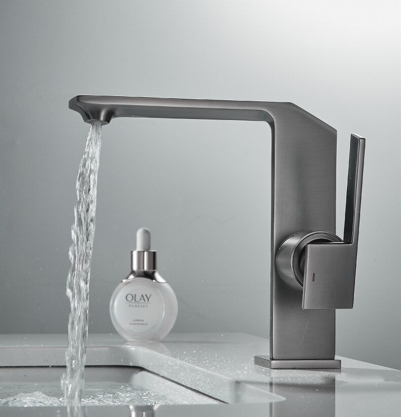 The Tiqui™ Solid Brass Bathroom Sink Faucet from Tiqui, featuring a modern matte gold finish and minimalist design, is pouring water into a white sink. It boasts a sleek, angular spout and an ergonomic single-lever handle, adding a touch of elegance to the bathroom with its sophisticated finish.