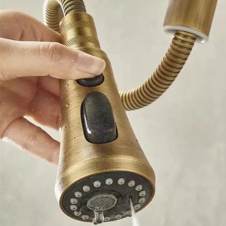 An Antique Brass Pull-Down Spout Kitchen Faucet by Rozin with a high arc and sleek, modern design pours water into a stainless-steel sink. The faucet handle is positioned to the right. In the background, reminiscent of elegant bathroom faucets, two white canisters with wooden lids are on a marble countertop.