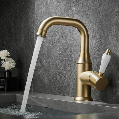 The Tiqui™ Single Handle Bathroom Sink Faucet, featuring a brass finish and white handle, is installed over a gray countertop, pouring a stream of water into the sink. The background features a dark textured wall and a small vase with white flowers.